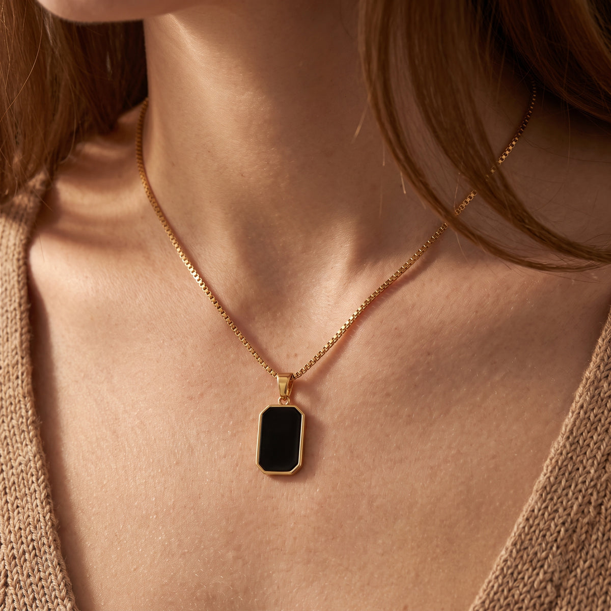 Close-up of a model wearing a gold-tone box chain necklace with an octagonal black pendant over a tan knit cardigan.
