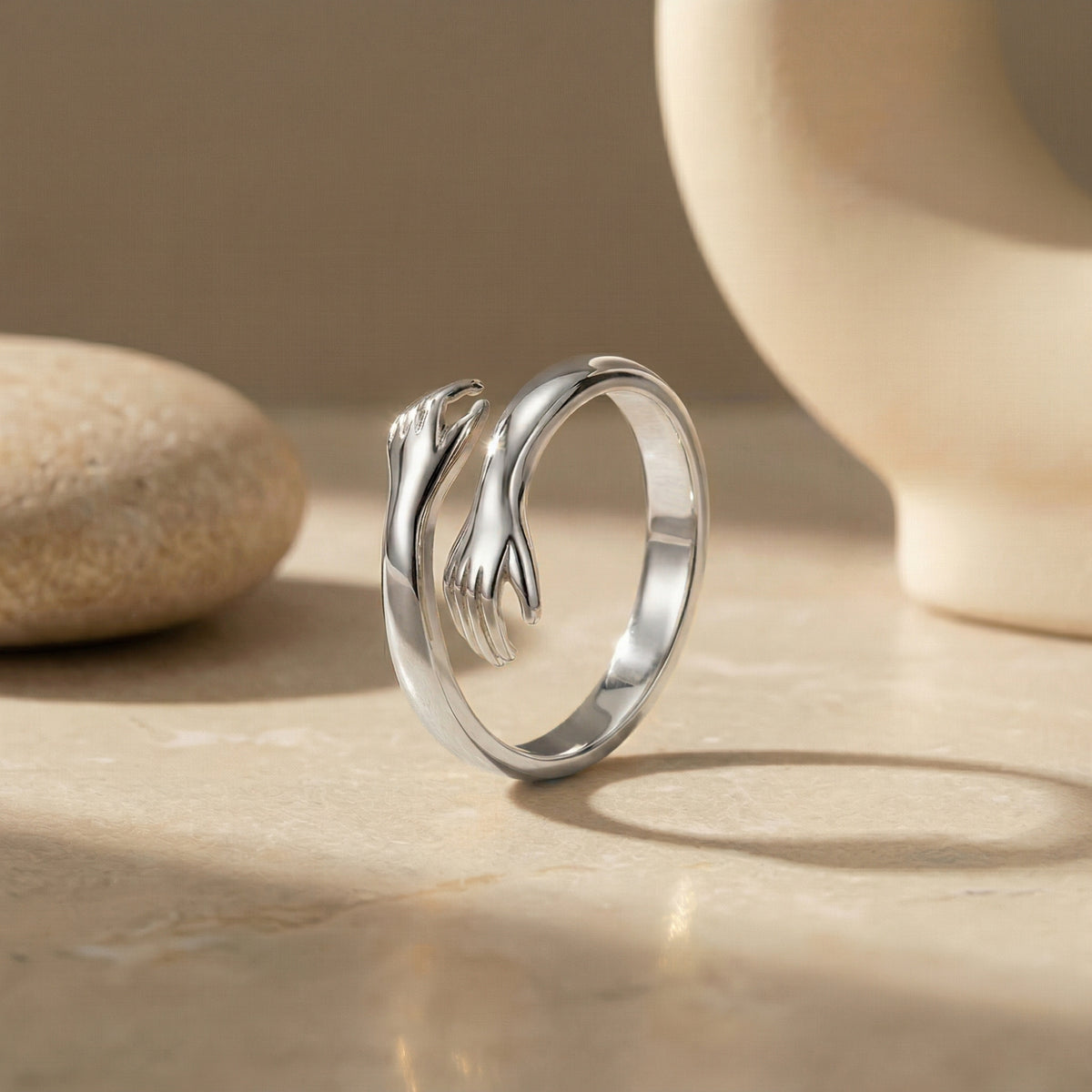 Silver-tone open-band hug ring with hugging hand detail standing upright on a beige surface.