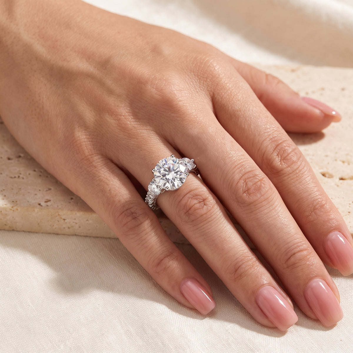 Alternate hand view displaying the round three-stone ring on a finger against a neutral textured backdrop.