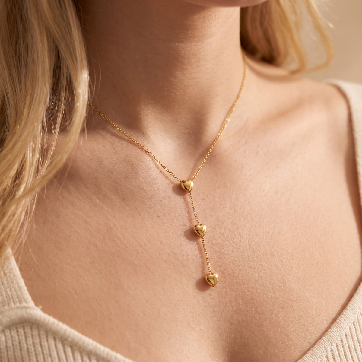 Angled close-up of a woman wearing a gold-tone triple heart lariat necklace over a ribbed knit neckline