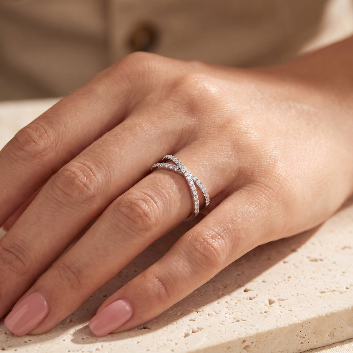 Silver crossover ring on a hand resting over a beige textured stone surface.