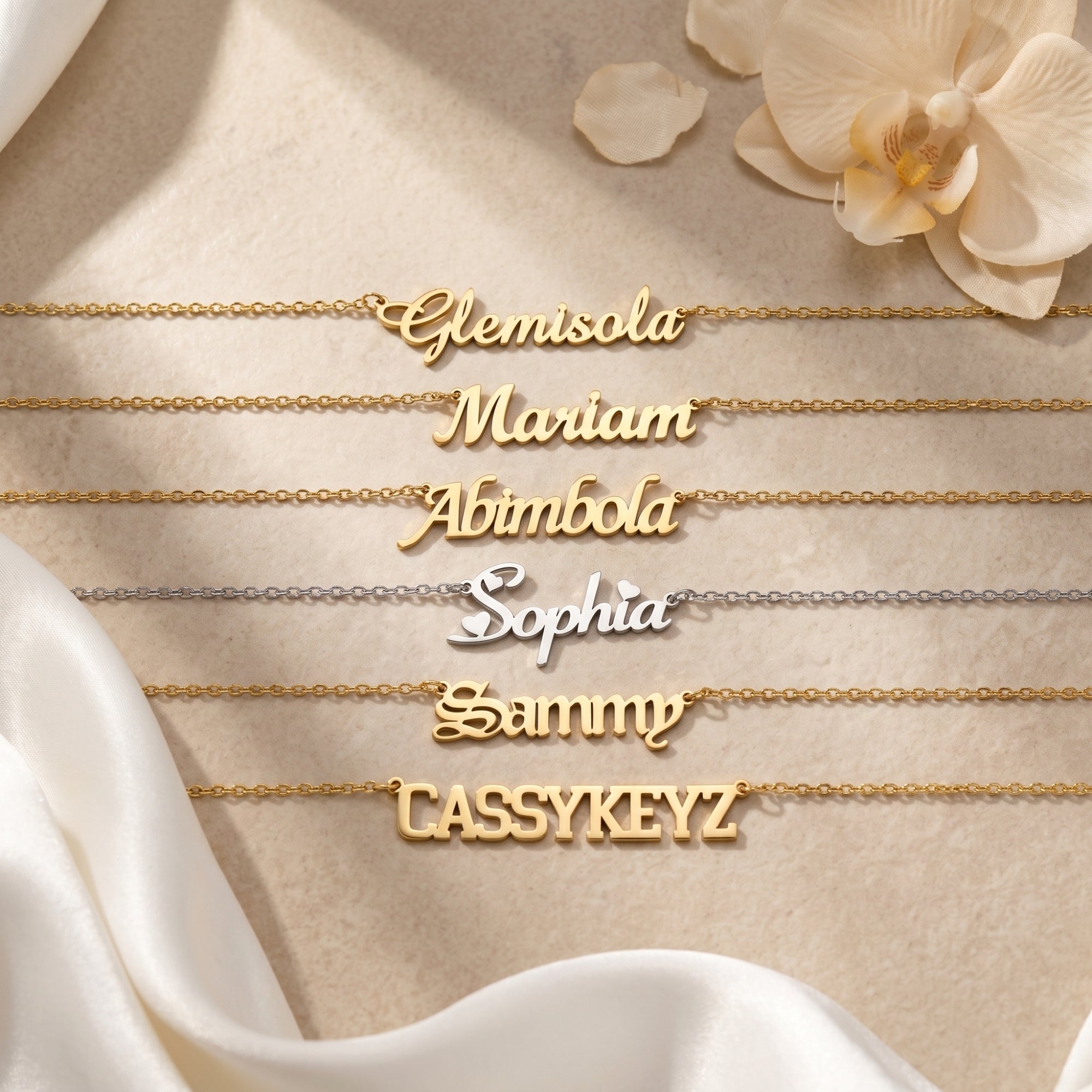 Personalized name necklaces in 18K gold and silver finishes displayed on a neutral surface with white orchids.