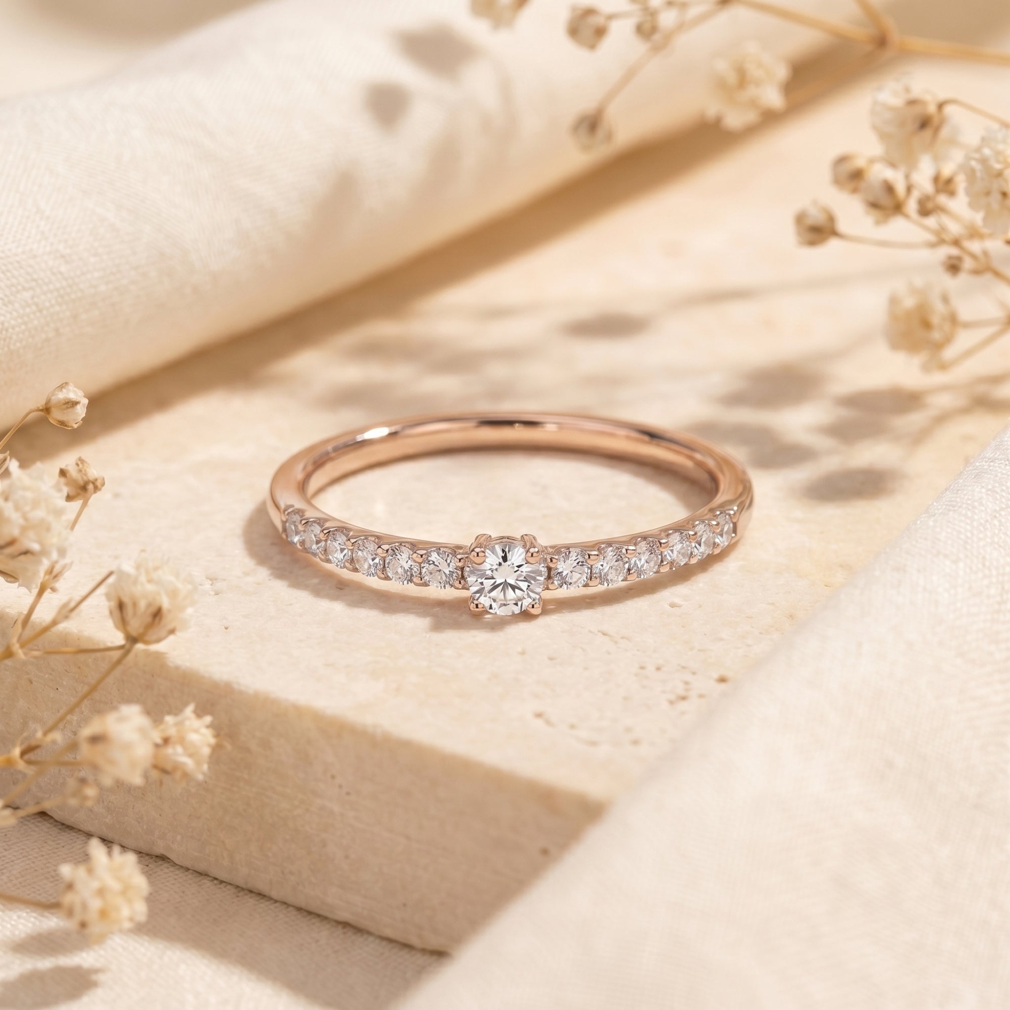 Rose gold round moissanite ring with pavé band on a textured stone surface with flowers.