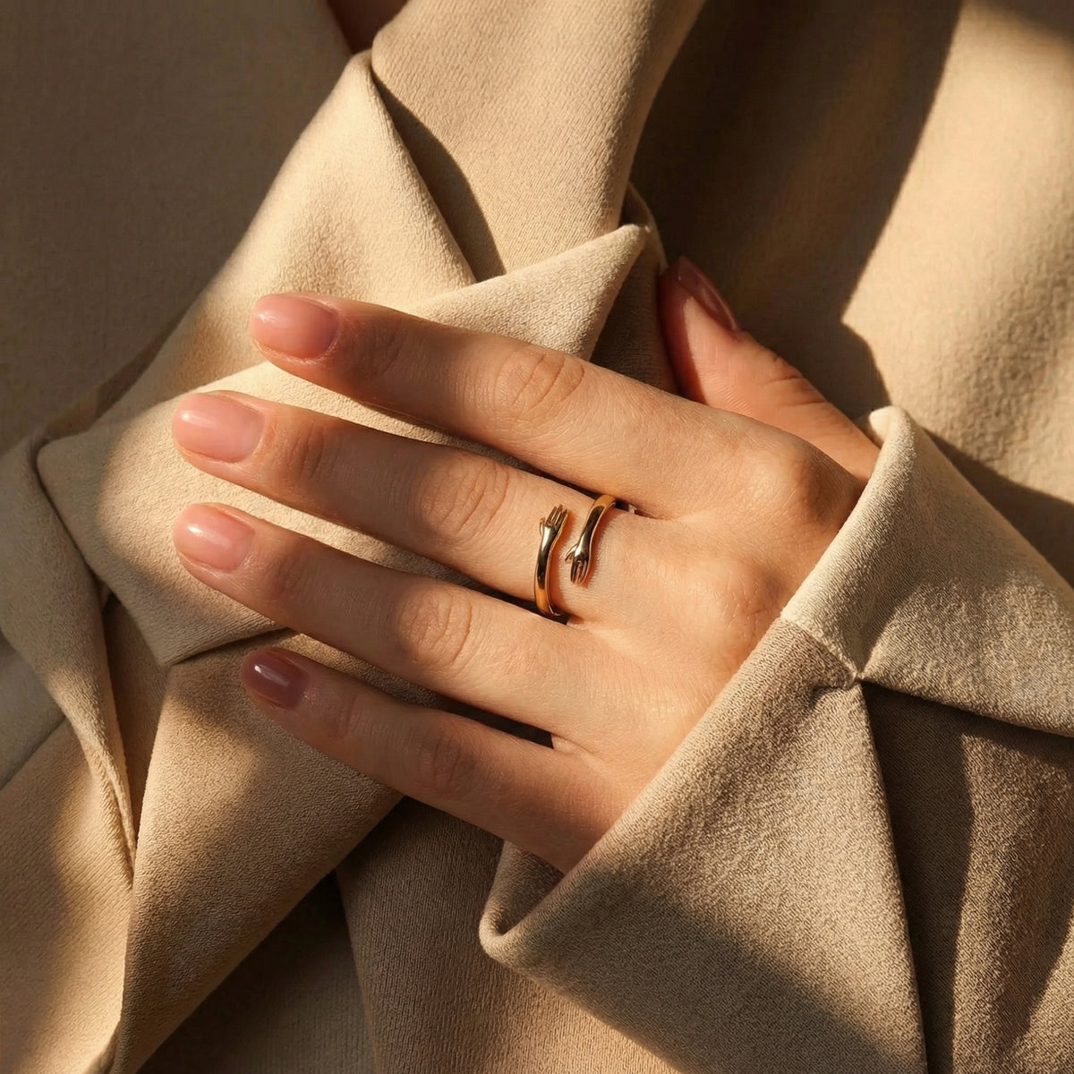 Gold-tone hug ring on a woman's hand resting over draped beige fabric.