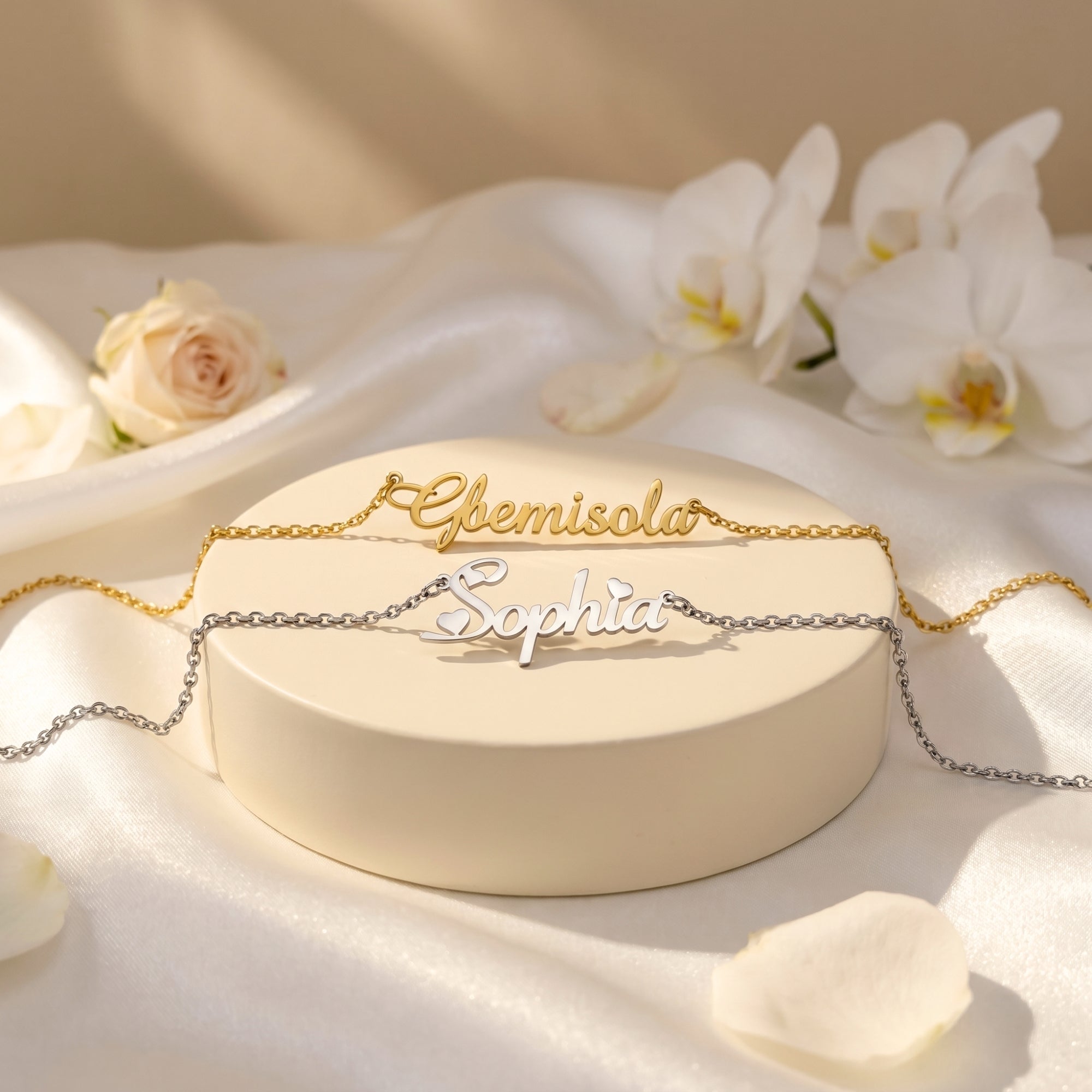 Two custom name necklaces in 18K gold and silver finishes on a round display stand with white silk and roses.