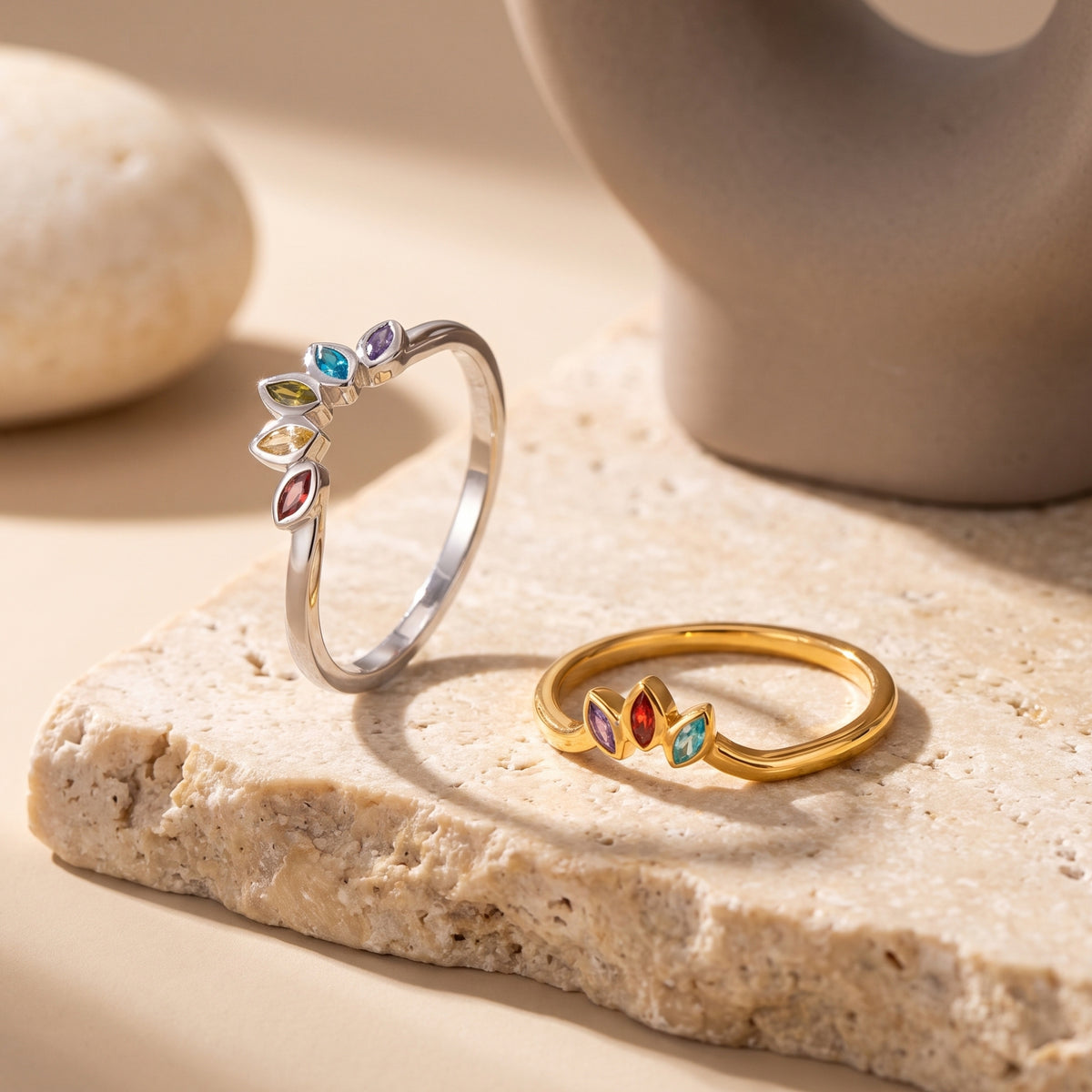 Silver five-stone and gold three-stone marquise rings displayed on a beige stone slab.