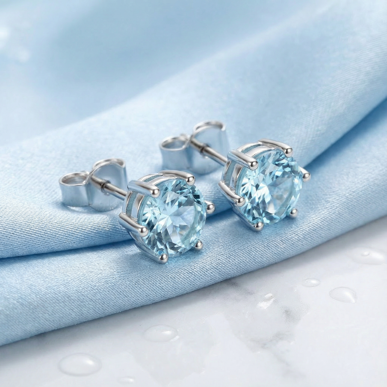 Round cut simulated aquamarine stud earrings in S925 sterling silver resting on light blue draped fabric.