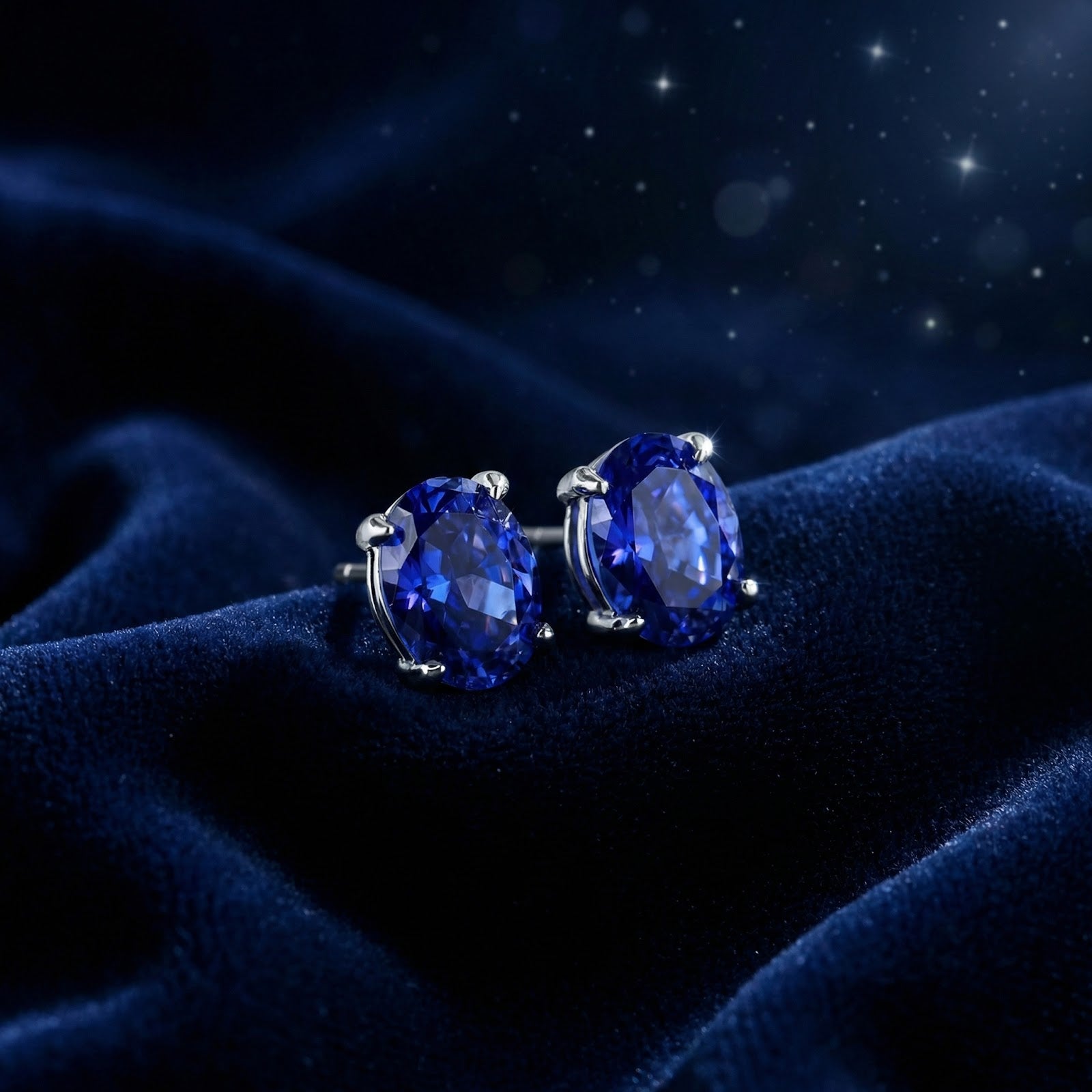 Oval cut simulated sapphire stud earrings in sterling silver resting on dark blue velvet fabric.