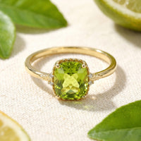 Cushion cut simulated peridot ring in solid gold on a textured surface with citrus leaves.