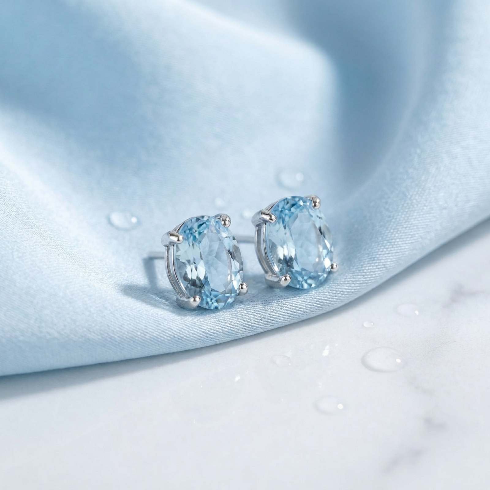 Oval cut simulated aquamarine stud earrings in sterling silver on light blue silk fabric with water droplets.