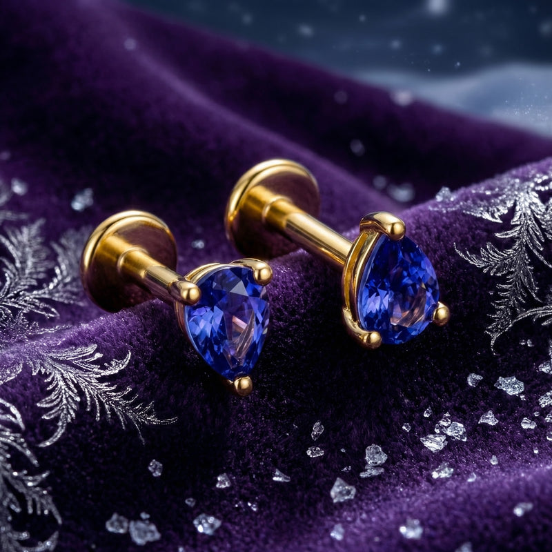 Close-up of pear-cut simulated tanzanite stud earrings in 18K gold vermeil resting on textured purple fabric.
