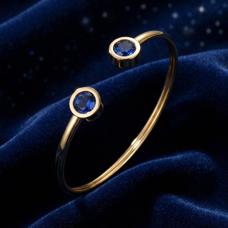Gold stainless steel Verian bangle featuring two round blue simulated sapphires on blue velvet.