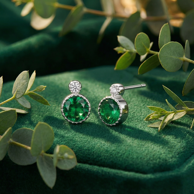 Round simulated emerald halo earrings in S925 sterling silver displayed on a green velvet surface with leaves.