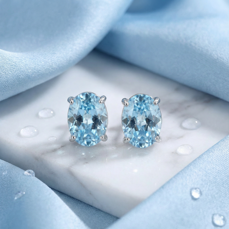 Pair of oval simulated aquamarine sterling silver stud earrings displayed on a white marble base with blue silk.