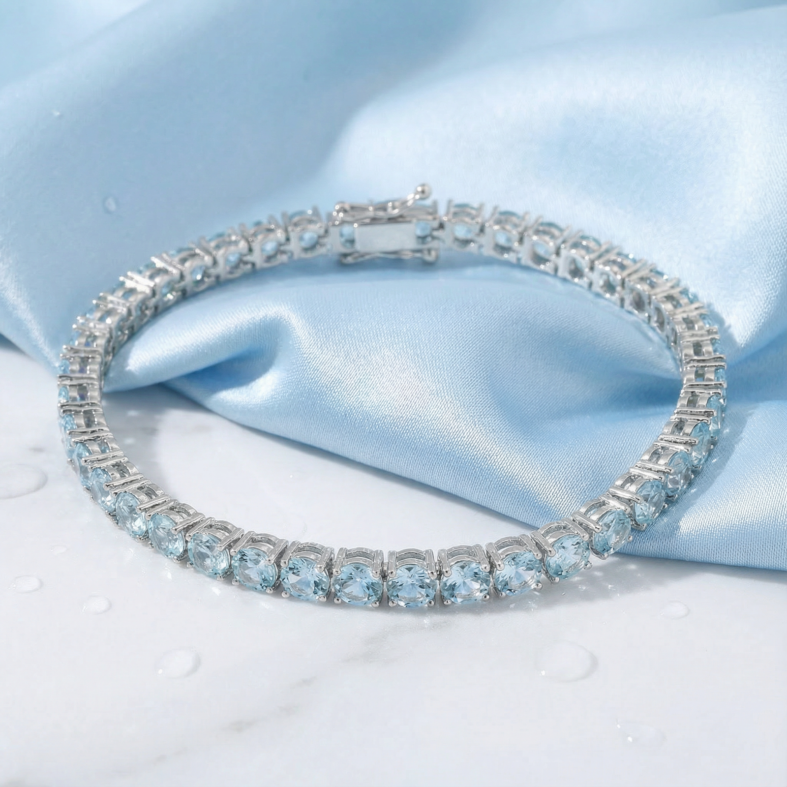 Round cut simulated aquamarine tennis bracelet in stainless steel on light blue silk with water droplets.