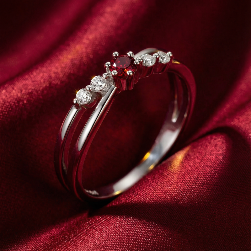 Angled view of the round simulated ruby crossover ring in sterling silver on red silk fabric.