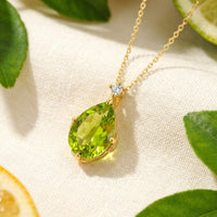 Pear-cut simulated peridot necklace in yellow gold on white fabric with citrus and green leaves.