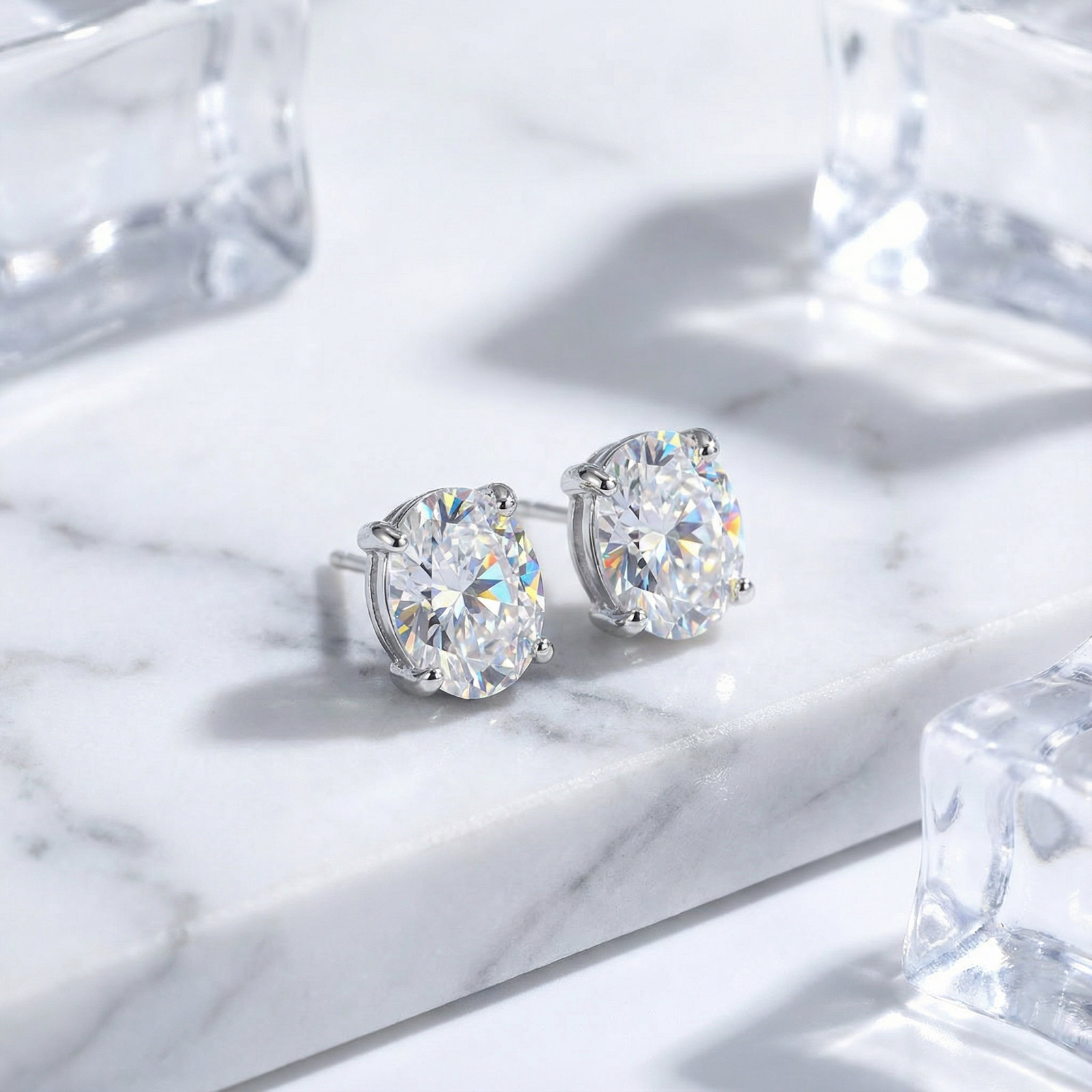 Oval cut zirconia stud earrings in S925 sterling silver settings on a white marble surface with ice props.