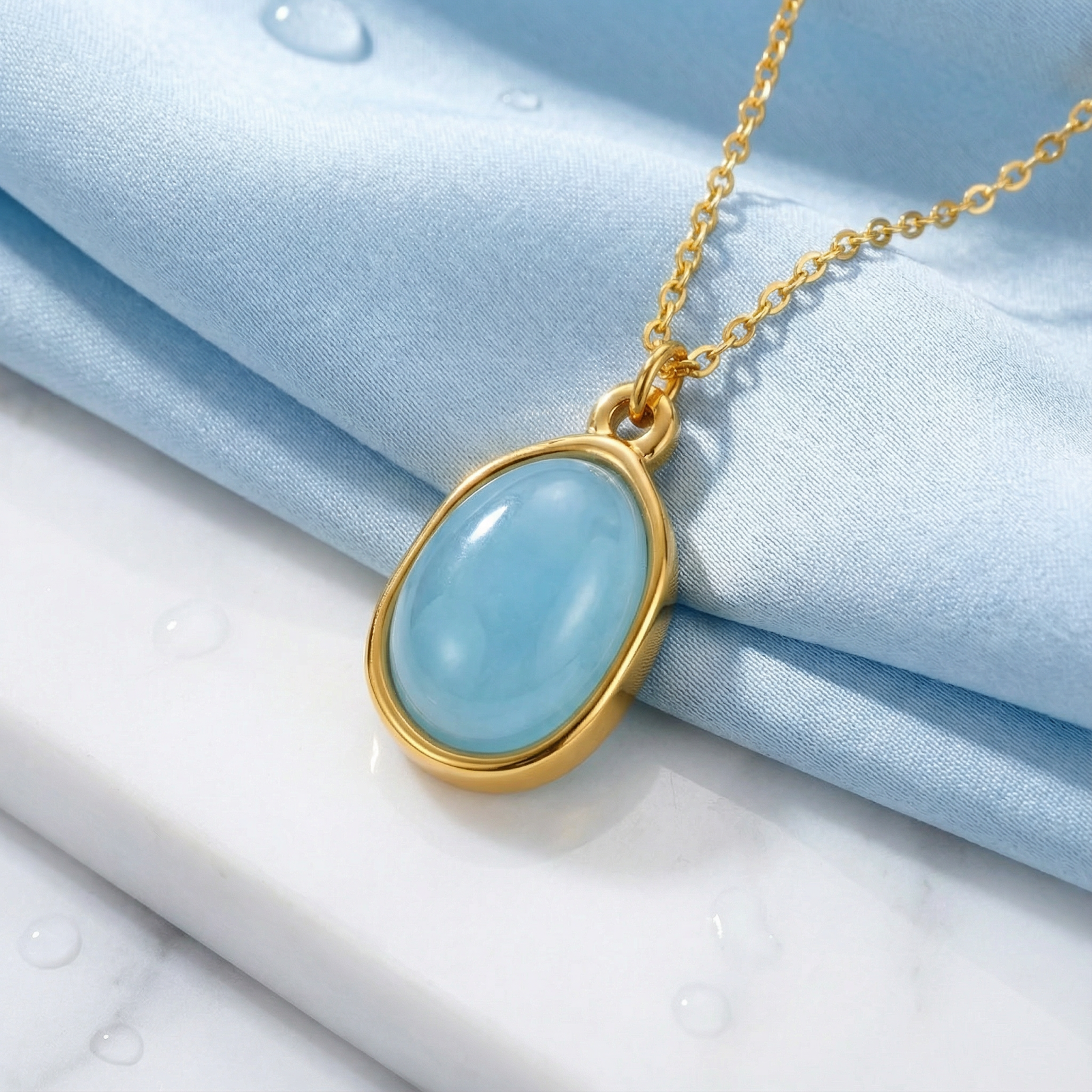Close-up of the gold bezel-set teardrop simulated aquamarine pendant on a gold chain over blue fabric.