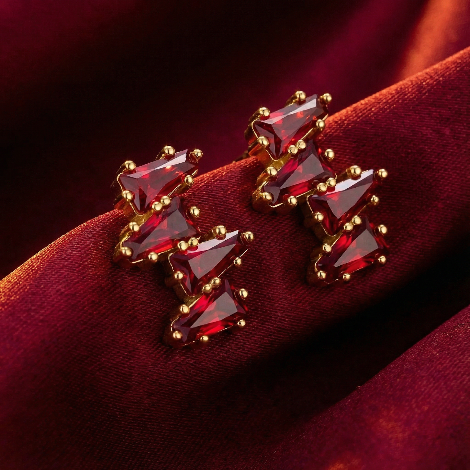 Angled view of Rhianna Studs in 18K gold vermeil with trillion-cut red rubies on a red silk background.