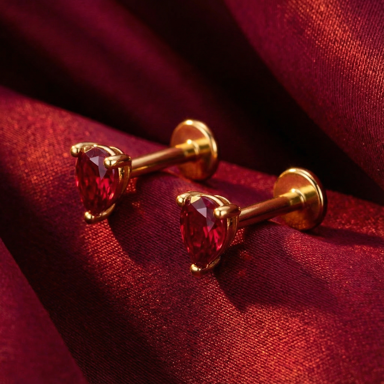 Pear cut simulated ruby stud earrings in 18K gold vermeil displayed on red silk fabric.
