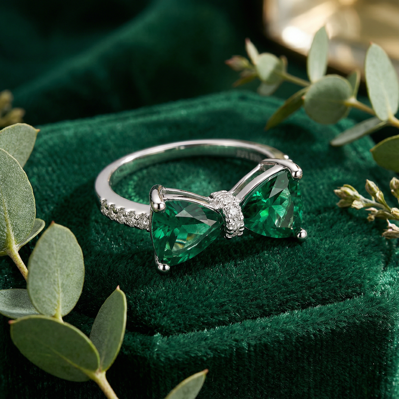 Close-up of a sterling silver bow ring featuring two trillion-cut simulated emeralds on a green velvet box.