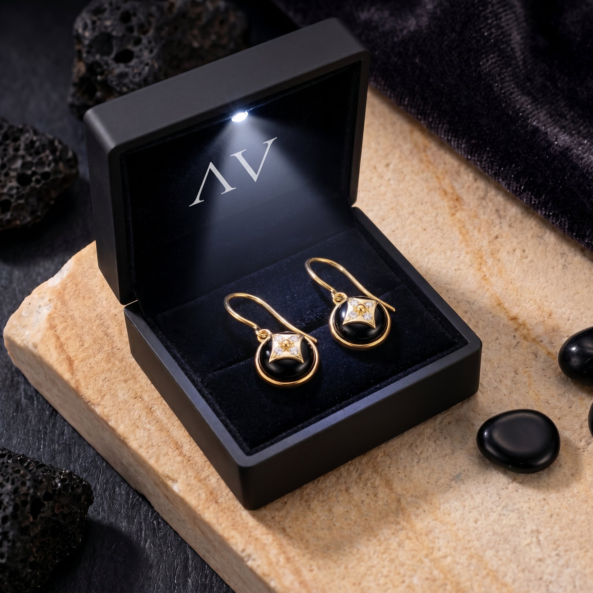 Yellow gold black onyx drop earrings presented in a black illuminated jewelry box on a stone surface.