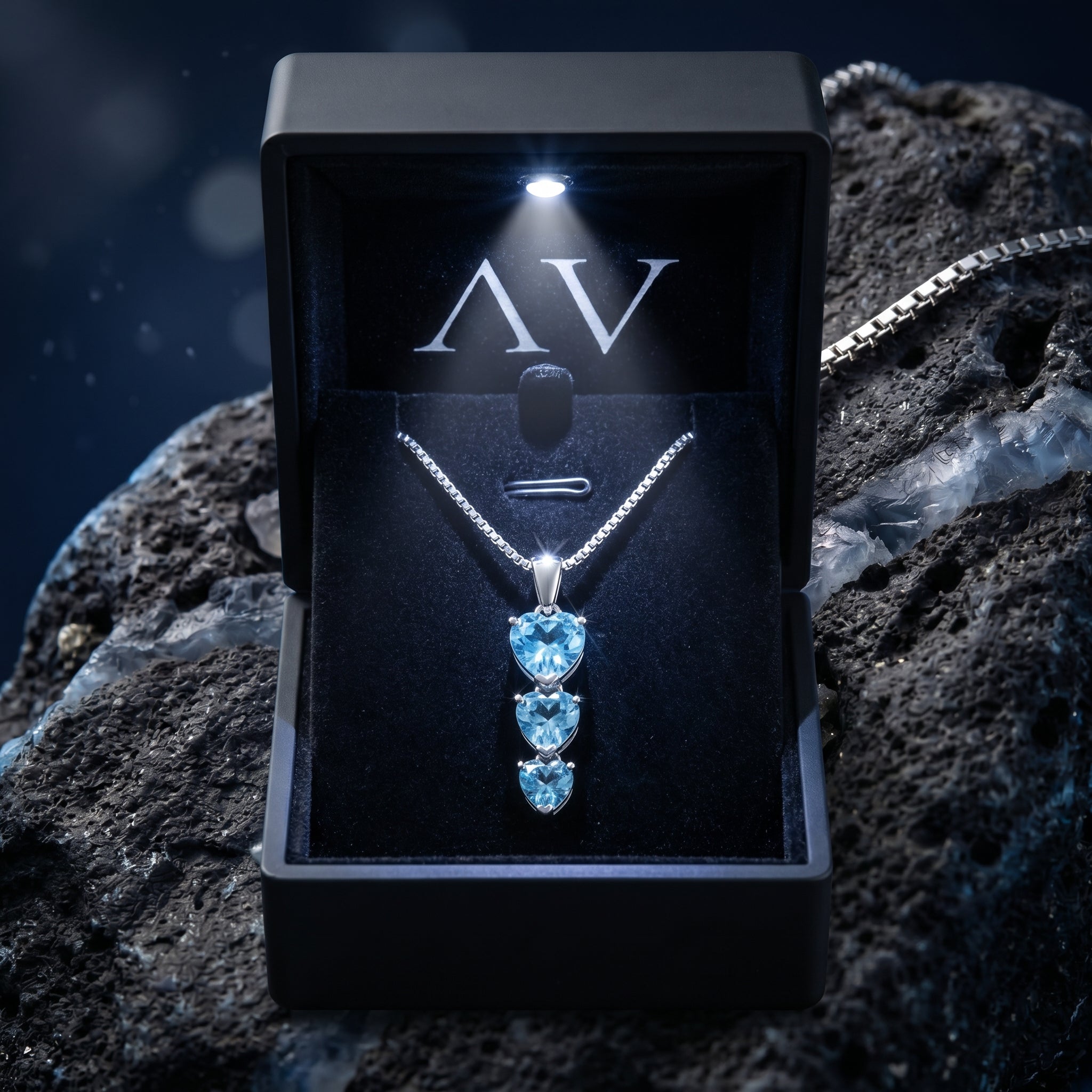 Silver heart cut blue topaz necklace in a lighted black jewelry gift box on a dark rock.