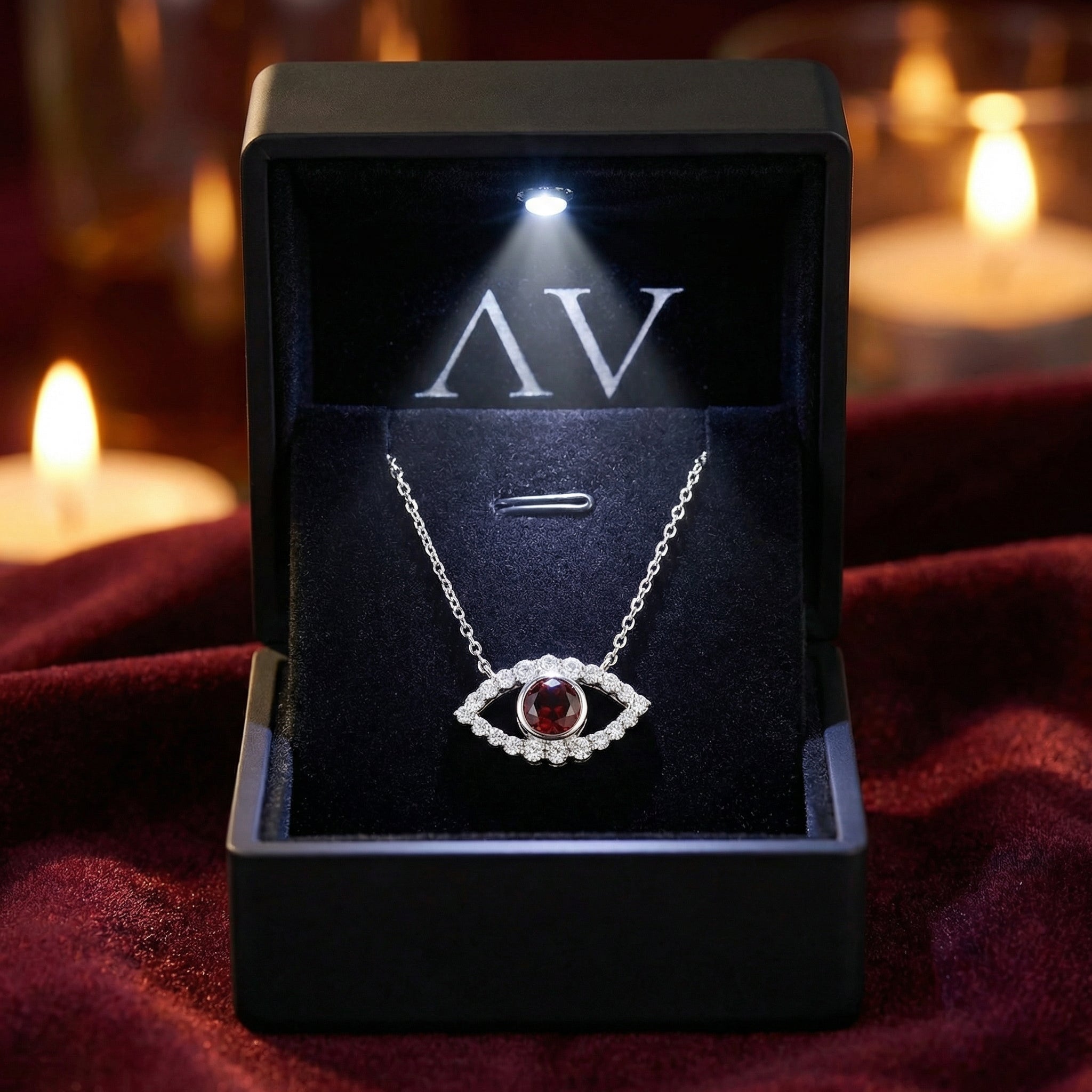 Sterling silver eye necklace with red garnet in a lighted black jewelry gift box on a red velvet surface.