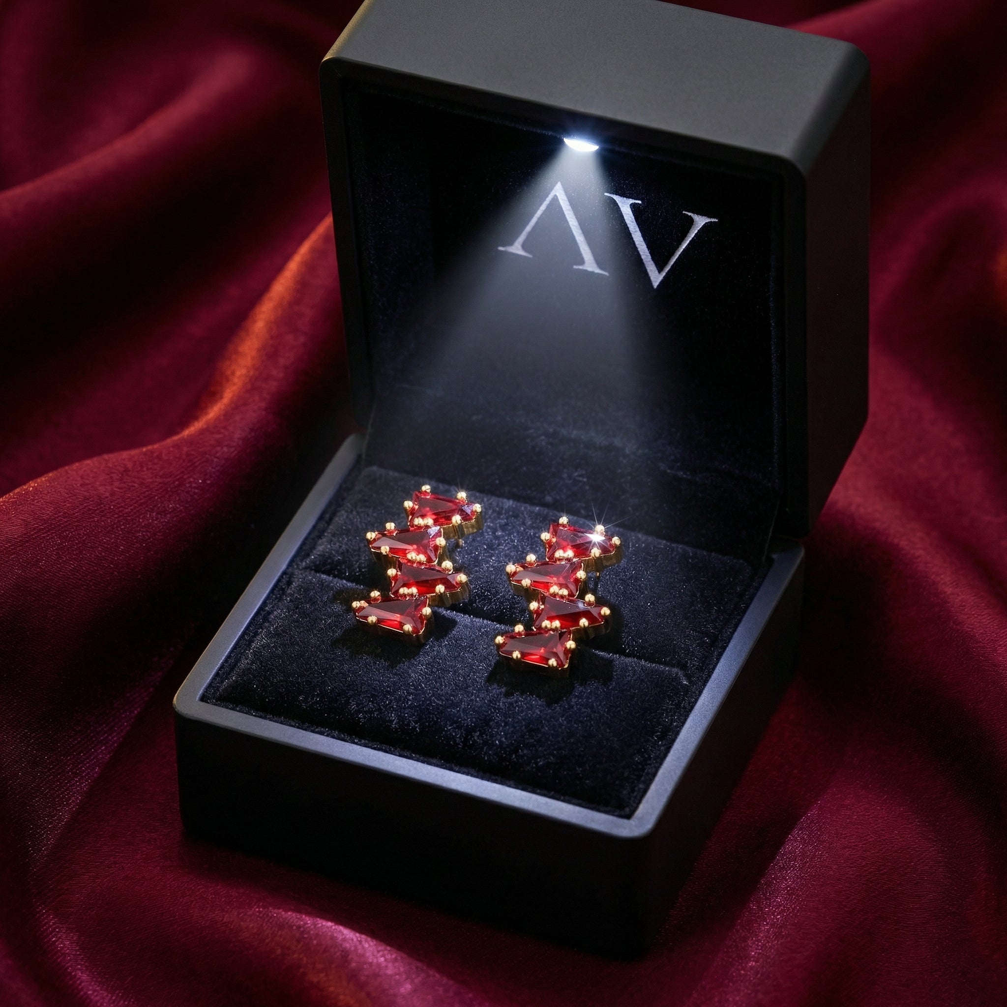Rhianna Studs with red trillion-cut rubies in a black LED-lit jewelry box on a red fabric background.