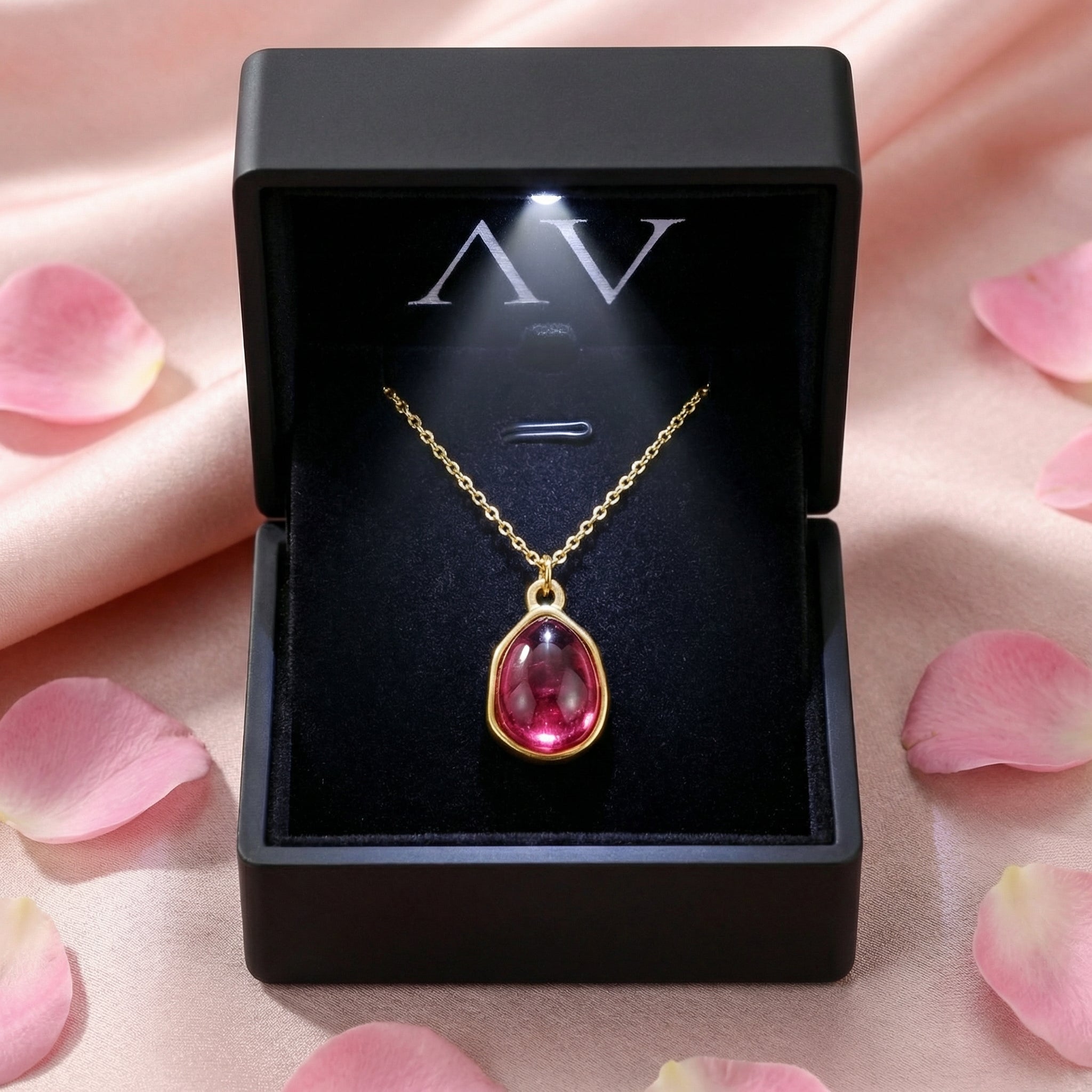 Gold teardrop pink tourmaline necklace inside a black lighted jewelry box surrounded by rose petals.