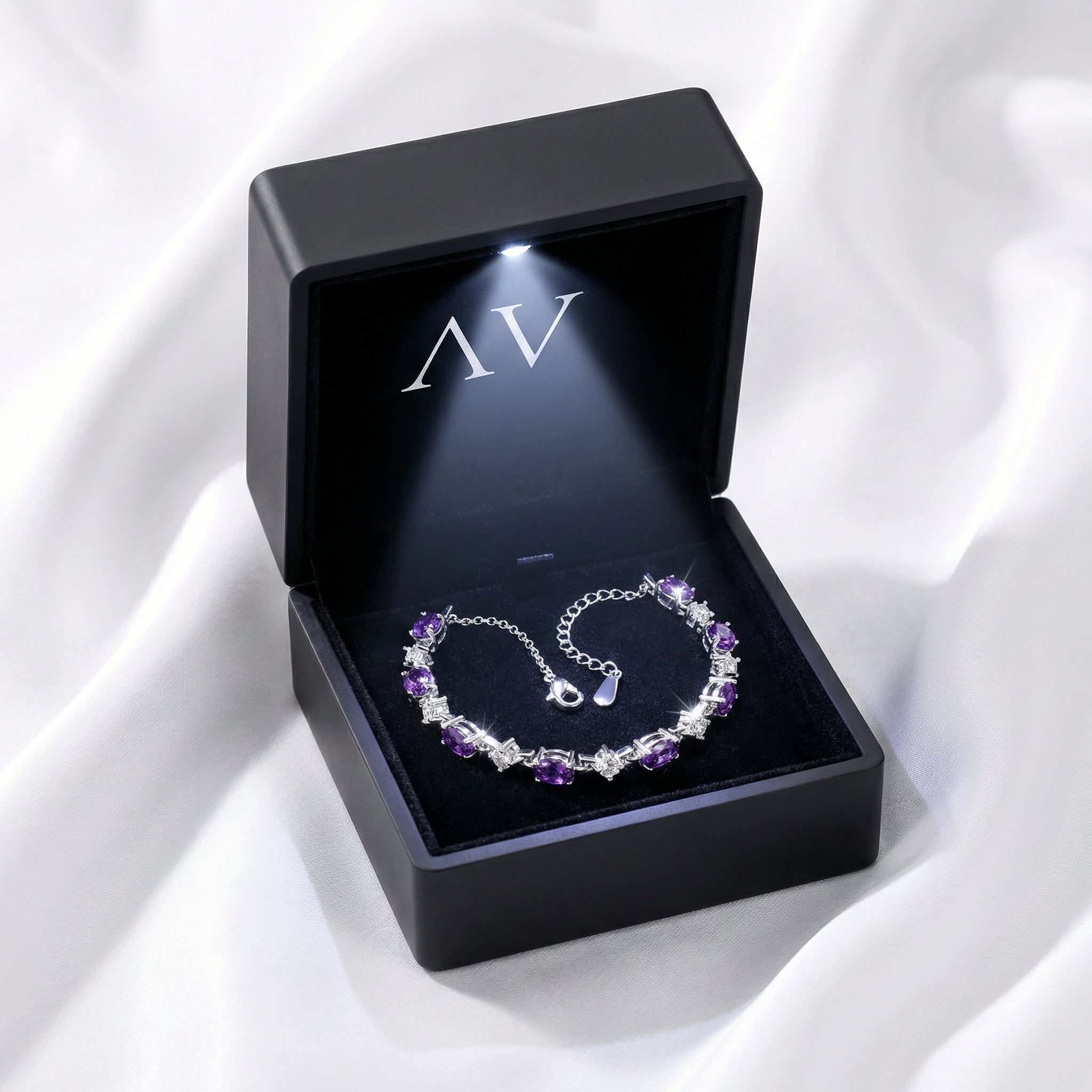 Wide view of the Lucina amethyst tennis bracelet in a black LED-lighted presentation box on white fabric.