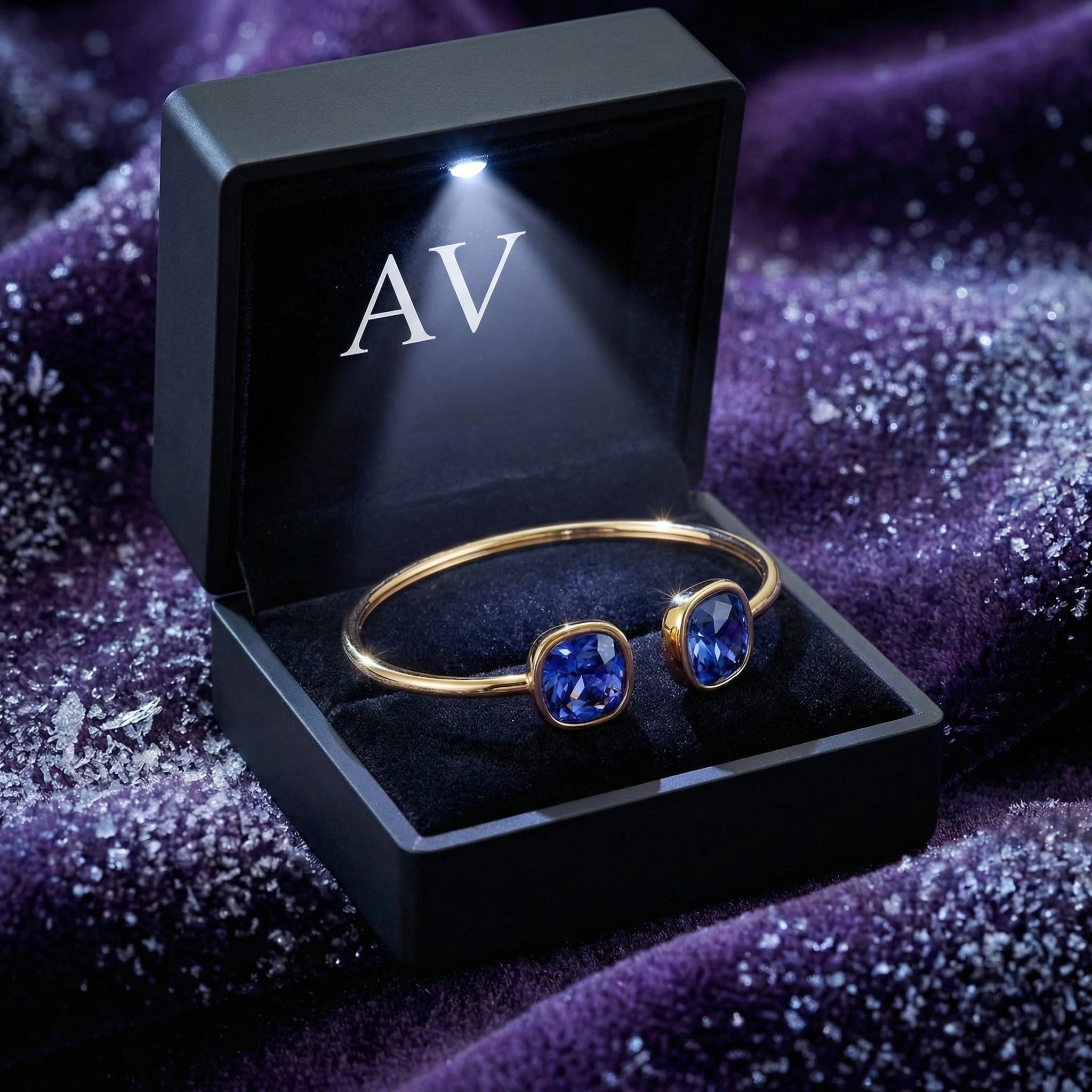 Gold Nerina bangle with simulated tanzanite stones inside a lighted black gift box on purple velvet.