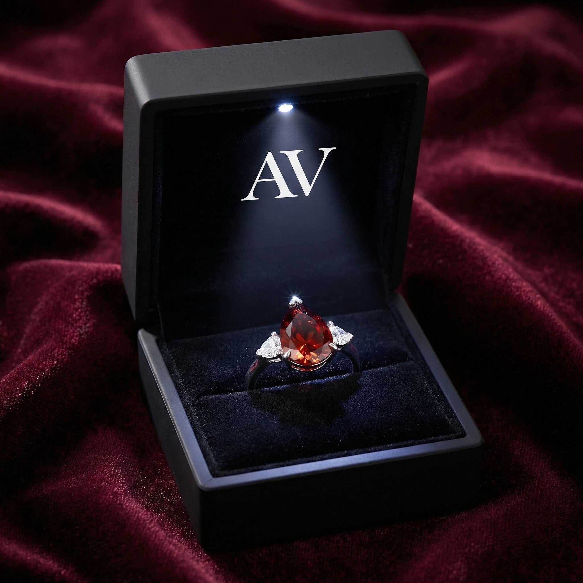 Pear-cut red garnet ring in a lighted black presentation box with AV logo on a red velvet surface.