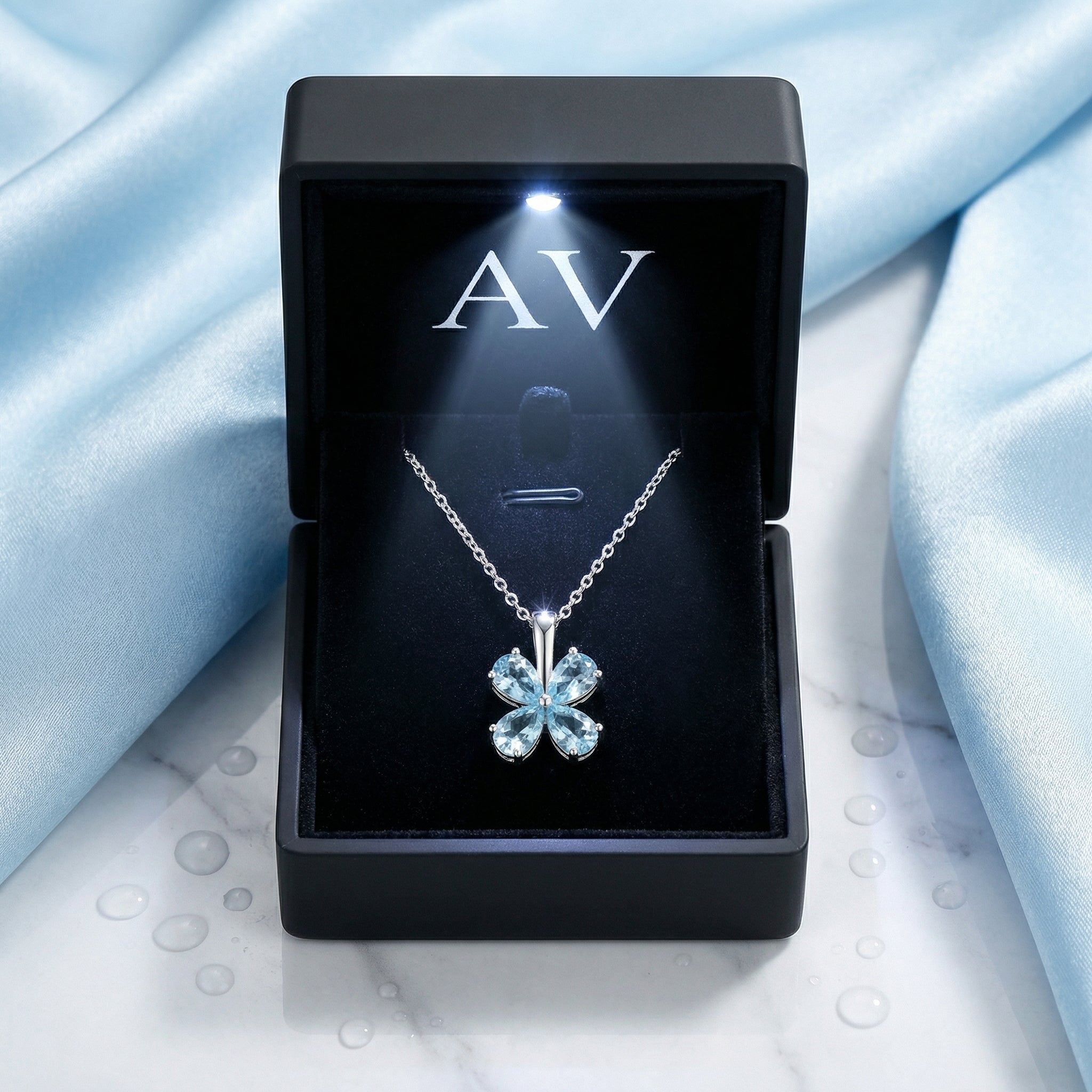 Silver clover necklace with blue simulated aquamarines in a black LED-lit gift box on blue silk and marble.