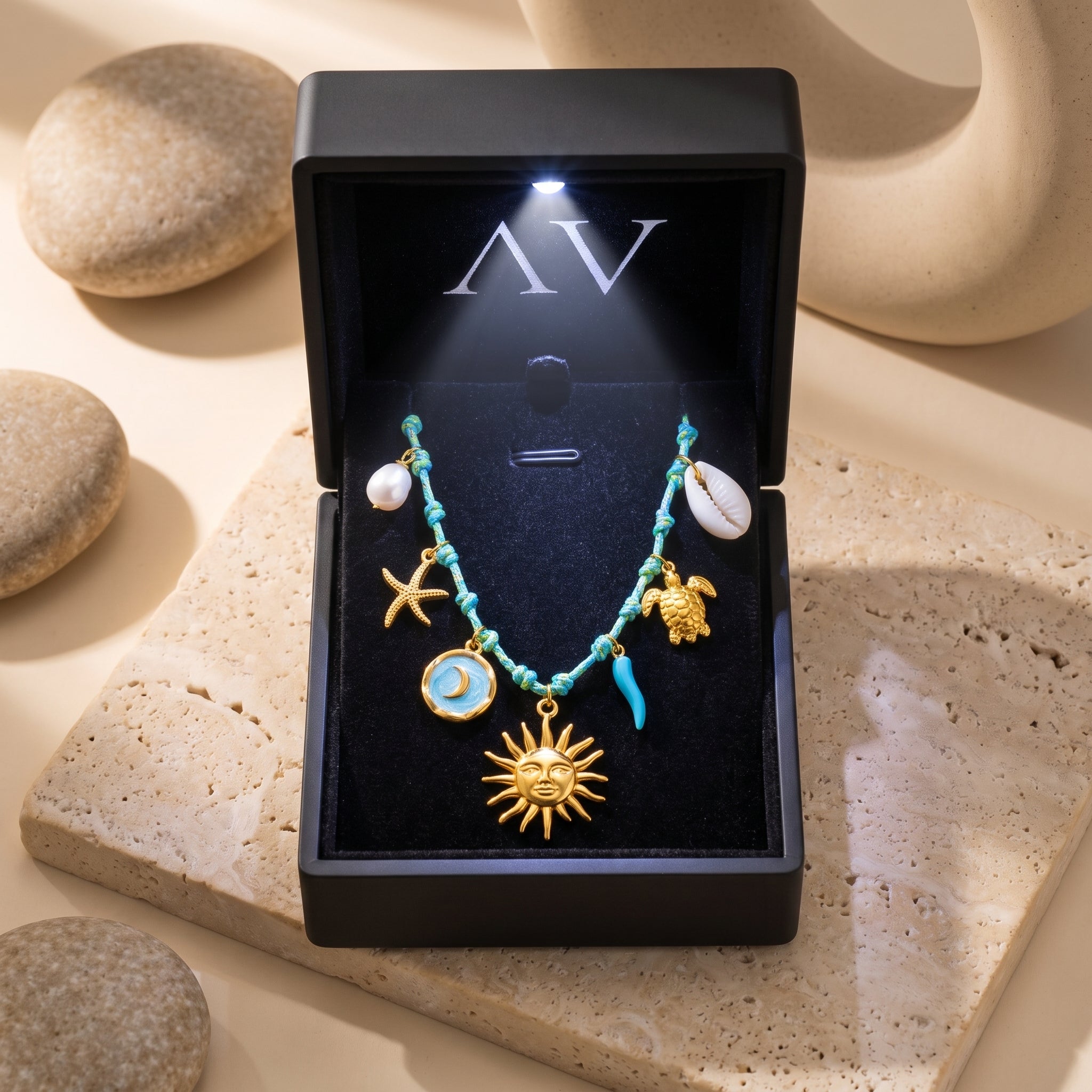 Gold charm necklace with sun and sea symbols displayed in a black LED-lit jewelry box on a stone surface.