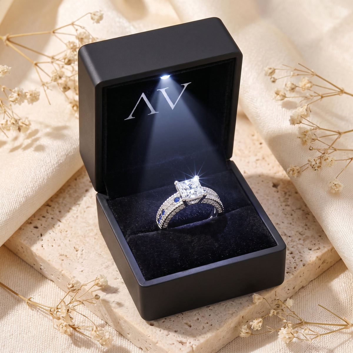 Princess-cut silver ring with blue accents inside a black LED-lit gift box.