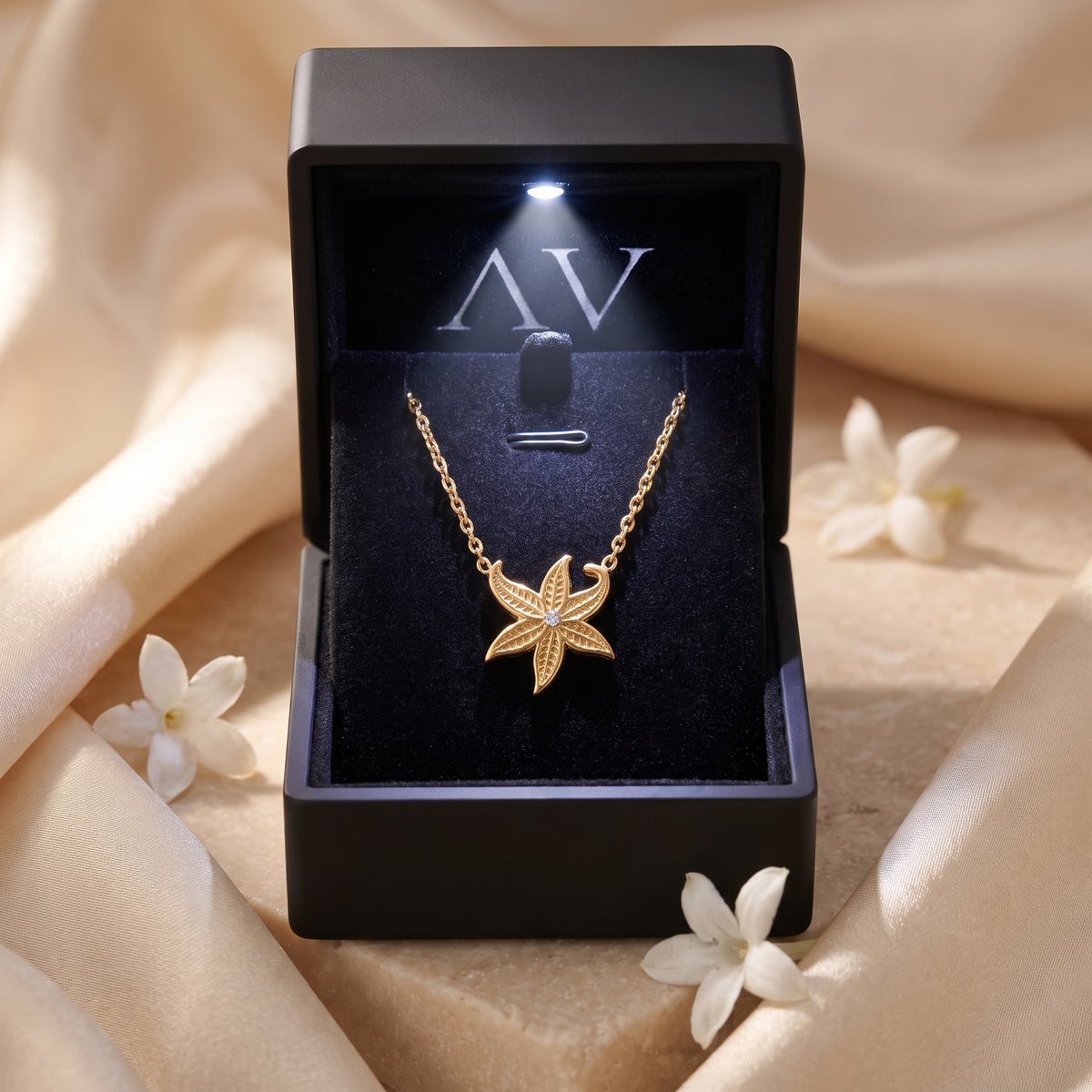 Gold starfish necklace inside a black LED-lit gift box with white flowers and silk background.