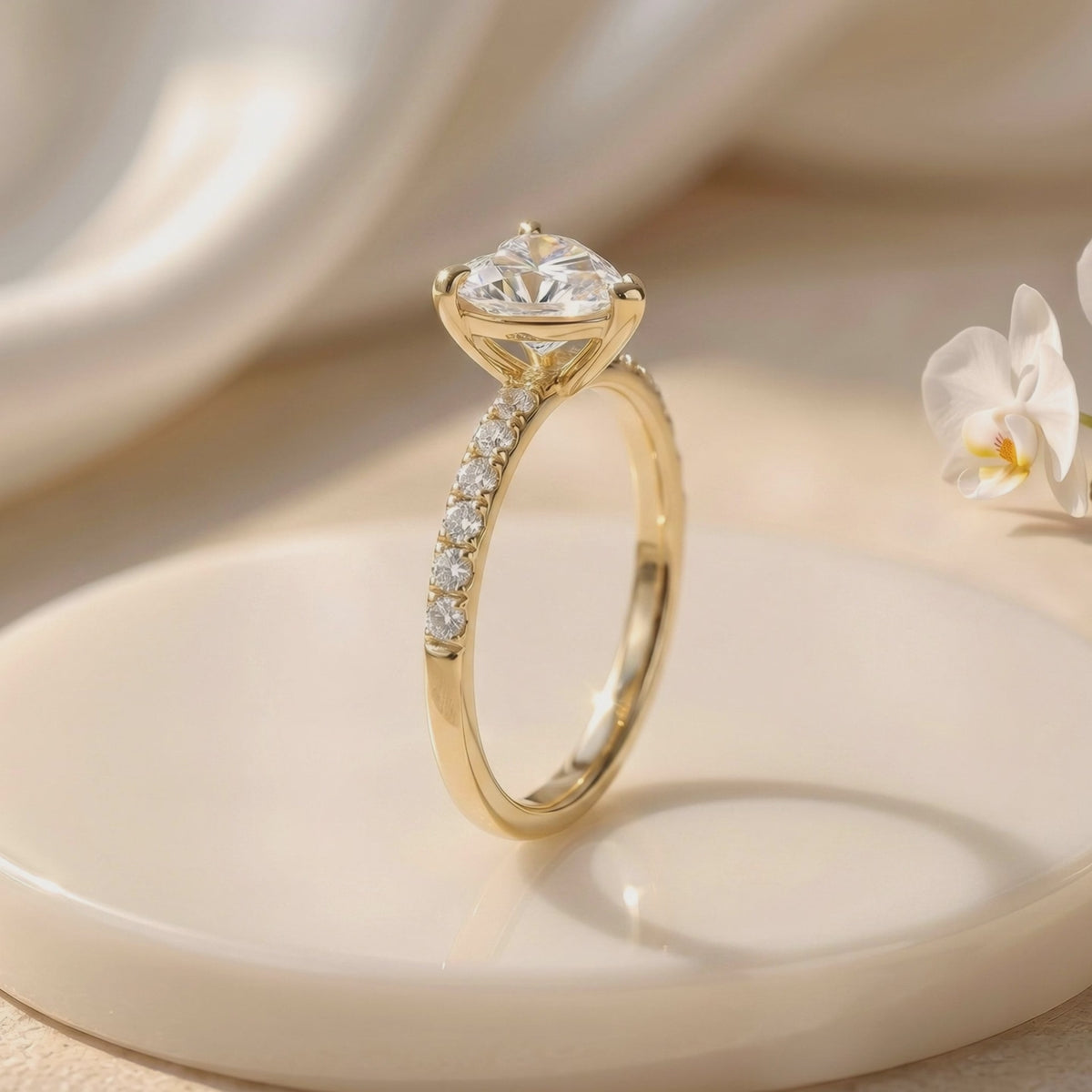 Side view of a gold-colored heart-cut solitaire ring with a pavé band on a white display platform beside an orchid.