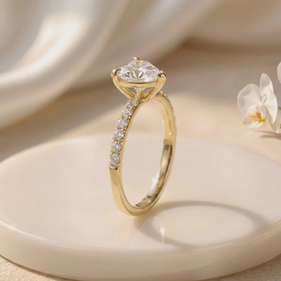 Side view of a gold-colored heart-cut solitaire ring with a pavé band on a white display platform beside an orchid.