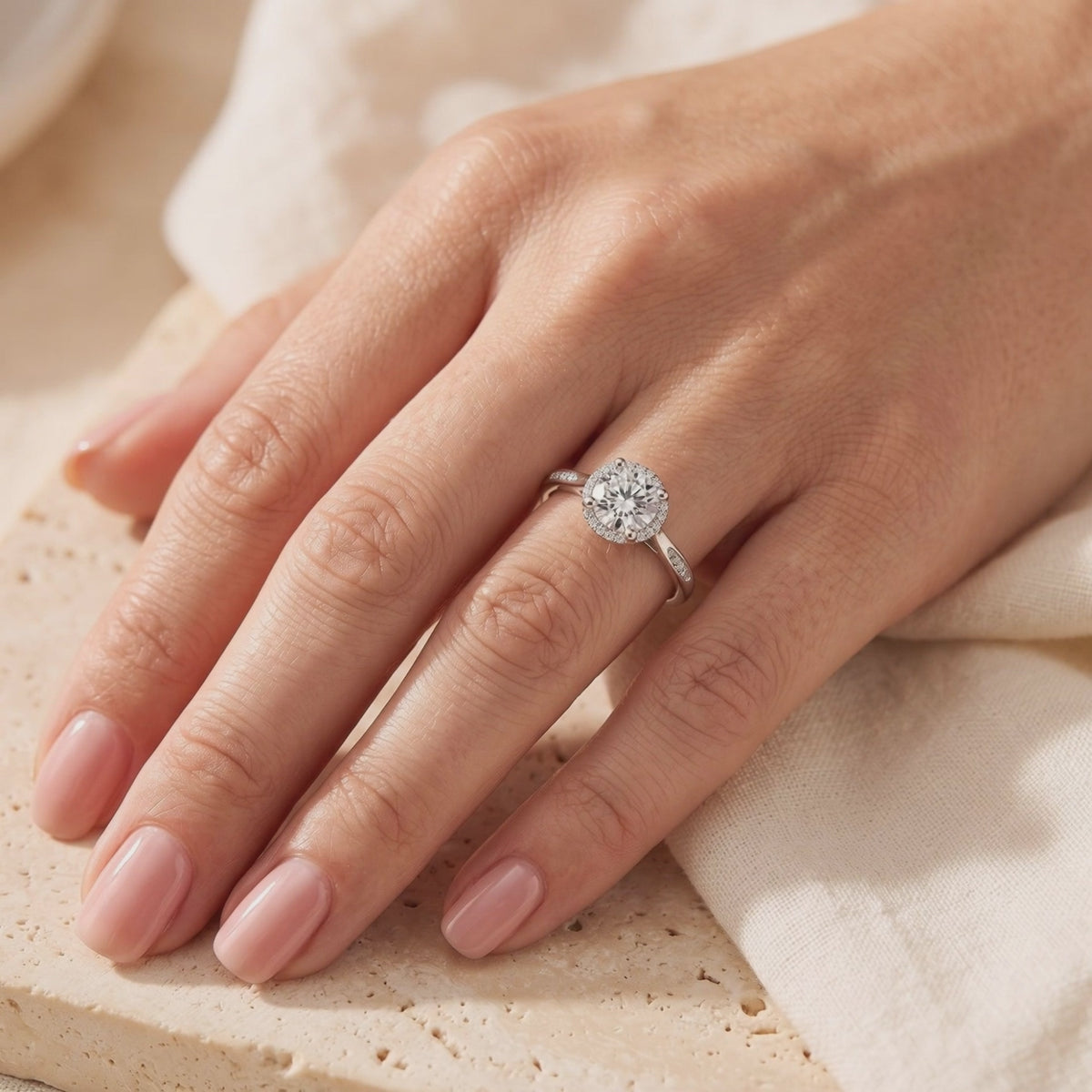 Silver round halo ring worn on a hand over a beige surface.