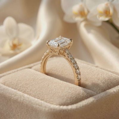 Side view of an emerald-cut solitaire ring with a pavé band resting in a beige velvet ring box.