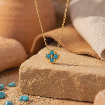 Gold chain necklace with a four-stone turquoise flower pendant displayed on a textured stone amid sand.