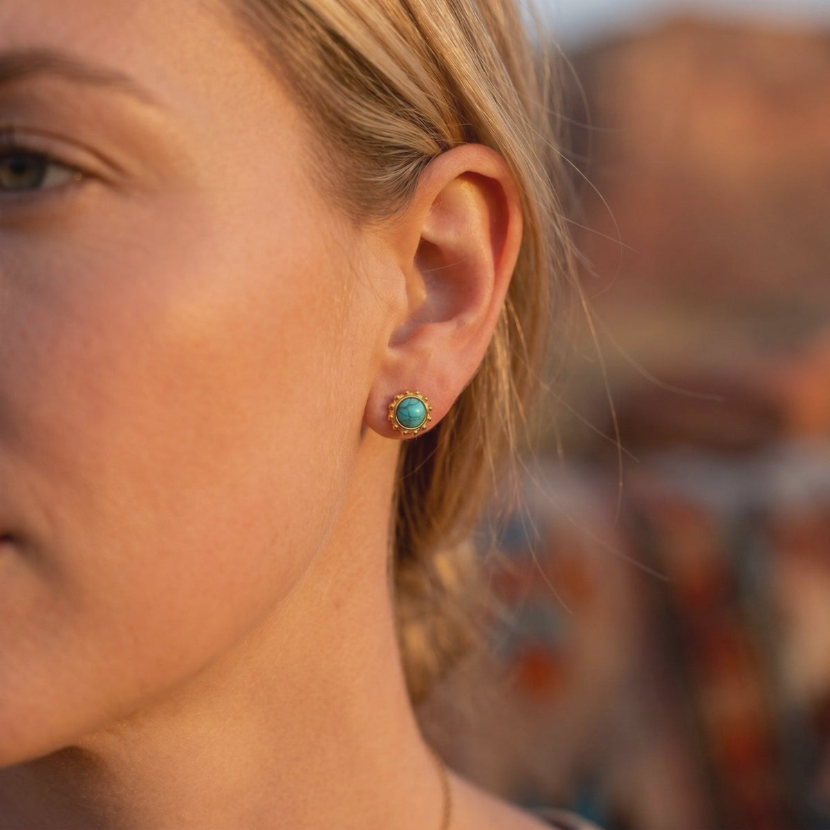 Close-up side view of a woman wearing a gold-tone turquoise-colored stud earring with a dotted bezel.