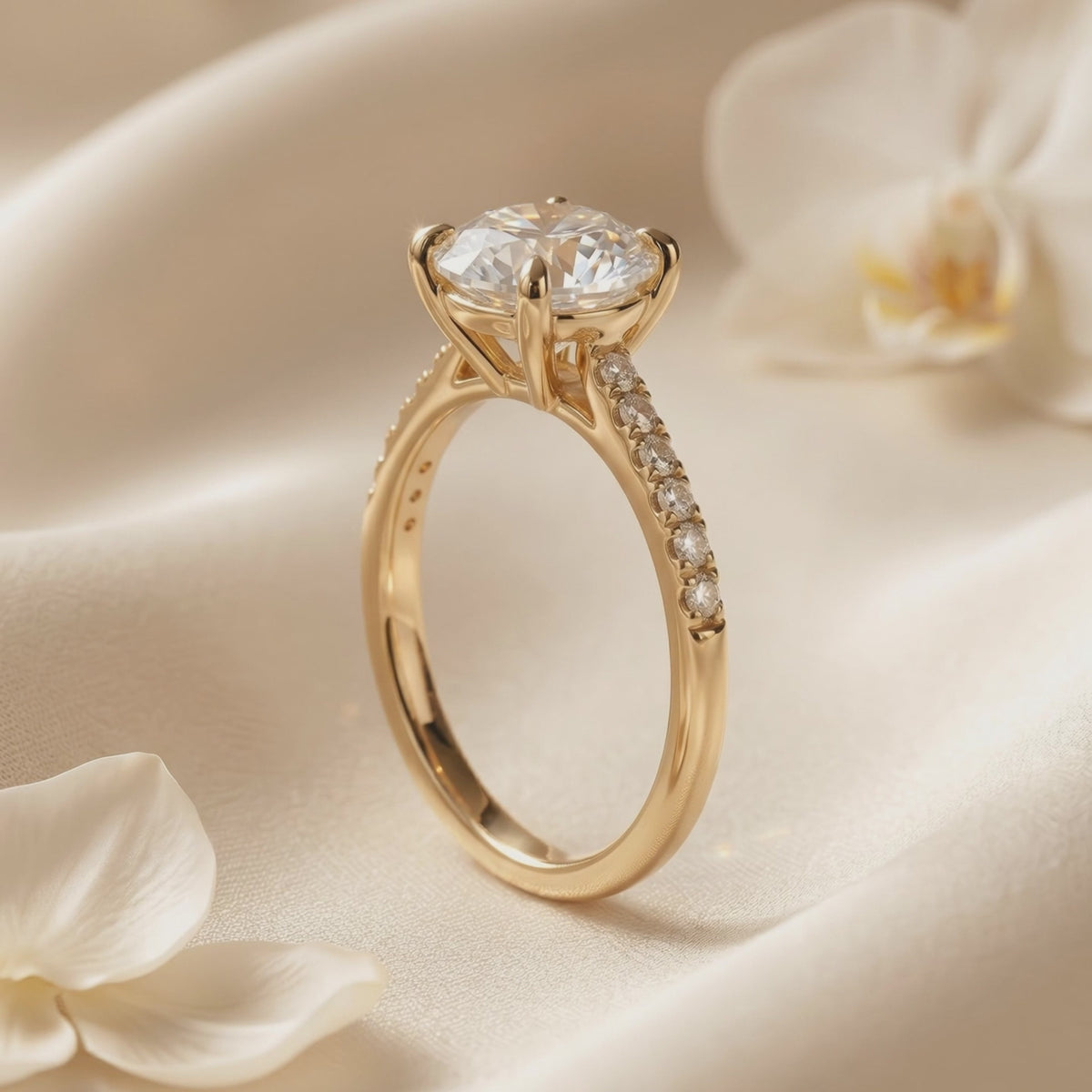 Gold-tone round solitaire ring shown upright in close-up on cream fabric beside white flower petals.