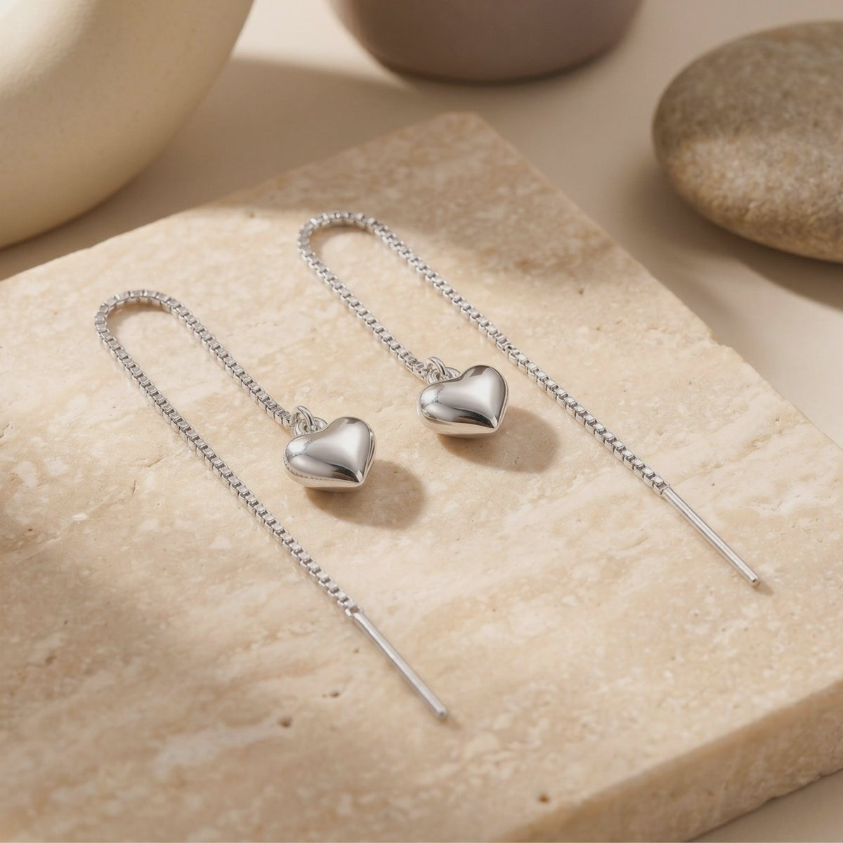Pair of silver-toned box-chain heart threader earrings arranged on a textured beige stone surface.