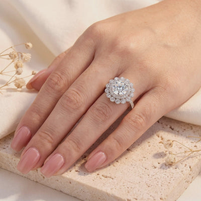 Silver floral halo ring with a round center stone, worn on a hand over a textured beige tile.