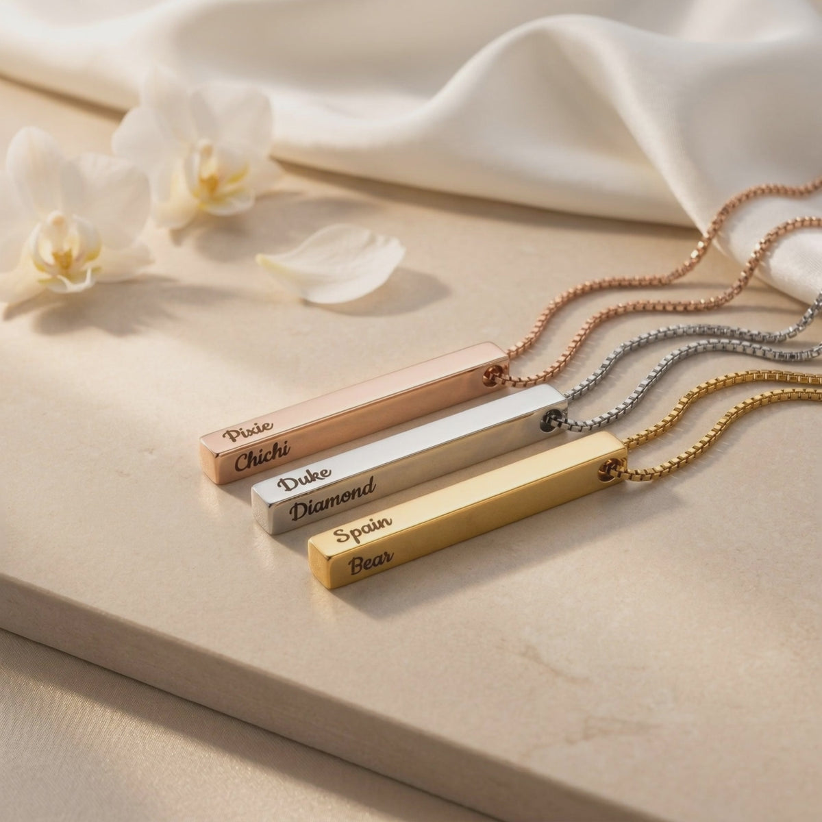 Three bar pendant necklaces in rose-gold, silver, and gold tones lie on a display surface with engraved text.
