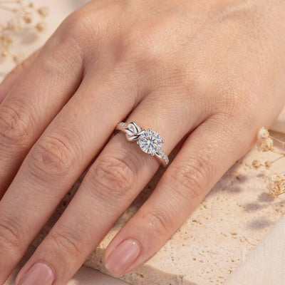 S925 sterling silver ring with a round clear center stone and love-knot detail, worn on a hand over a beige textured slab.