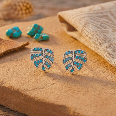 Pair of silver monstera leaf earrings with blue turquoise stones displayed on a tan wooden slab.